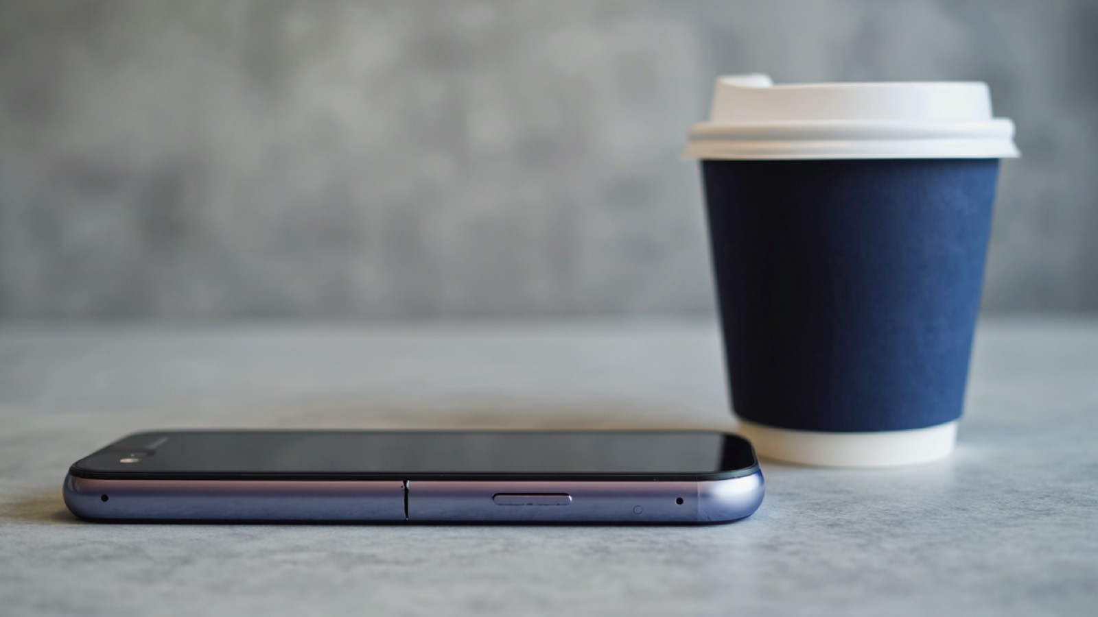 Ein Smartphone mit feinem Sprung im Display liegt umgedreht auf einer grauen Fläche neben einer schlichten Kaffeetasse